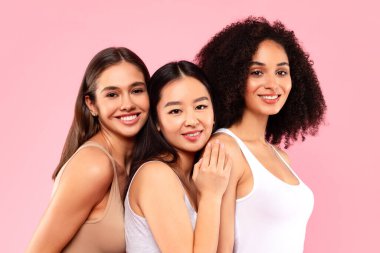 Pembe arka planda izole edilmiş kameraya poz veren ve gülümseyen genç uluslararası kadın arkadaşlar, stüdyo görüntüleri. Çeşitlilik, doğal güzellik ve moda.