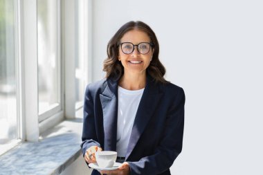 Mutlu, uzun saçlı, resmi kıyafet ve gözlük takan, elinde bardak tutan, kameraya gülümseyen, ofiste kahve molası veren, ofisini kopyalayan olgun iş kadını.