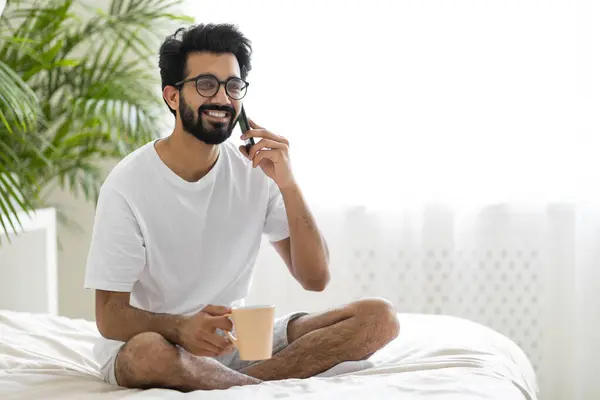 Home Leisure. Happy Young Indian Guy Sitting In Bed, Talking On Cellphone And Drinking Coffee, Smiling Male Having Pleasant Phone Call And Enjoying Hot Drink While Resting In Bedroom, Copy Space