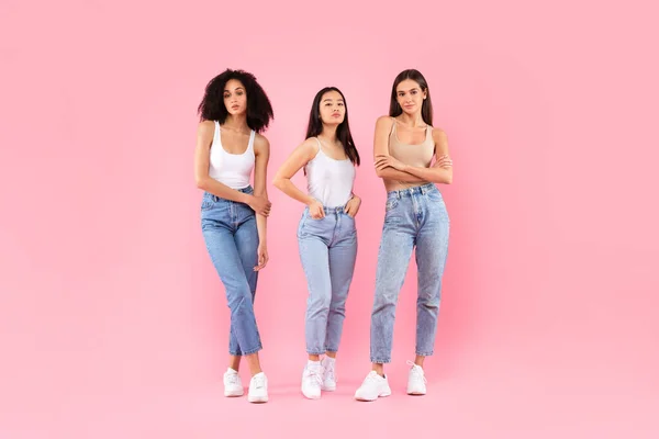 Three serious diverse women posing standing over pink studio background, full length shot of ladies friends trio looking at camera. Natural diverse female beauty
