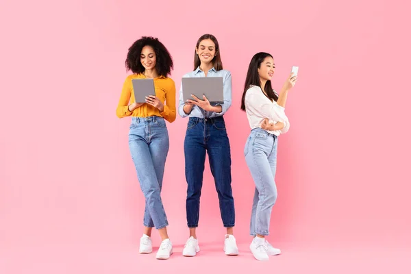 Happy multiracial ladies friends using different gadgets, using smartphone, laptop and digital tablet, networking online and browsing internet, standing on pink background, full length