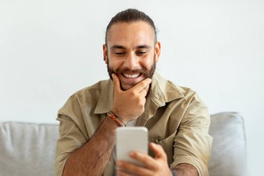 Evdeki kanepede oturan, telefon kullanan, sakalına dokunan, sevgilisiyle sohbet eden, haberleri kontrol eden, gündelik kıyafetler giyen yakışıklı beyaz adamın yakın çekimi. Güzel online teklif, anlaştık.