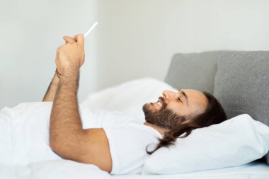 Sabahları beyaz tişört giyen, akıllı telefon kullanan ve gülümseyen, uyandıktan sonra kız arkadaşına aşk mesajları gönderen, beyaz tişörtlü mutlu ve uzun saçlı bir adamın yan görüntüsü.