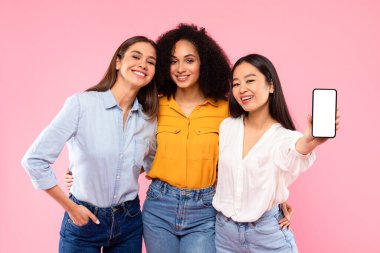 Tavsiye ediyoruz. Mutlu genç çok ırklı kadın arkadaşları cep telefonlarını beyaz ekranla gösteriyorlar. Model, reklam, uygulama veya web sitesi tasarımı, afiş, pembe stüdyo arka planı için.