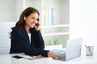 Resmi takım elbiseli, arkadaş canlısı, olgun bir kadın ofiste bilgisayar başında oturmuş kulağına kulaklık bastırıyor. İş kadınlarının online toplantısı var