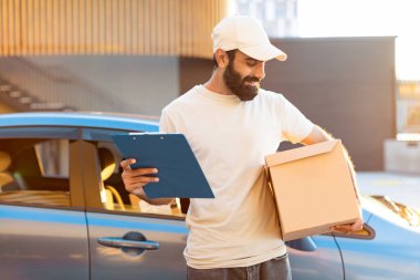 Paketler Nakliye Teklifi. Arabasının yanında duran Hint kuryesi, paket kutusu ve pano tabletini tutuyor, müşteri kapısına paket teslim ediyor, dışarıda poz veriyor. İşteki genç teslimatçının fotoğrafı.