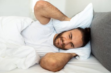Evde pijama giymiş sinirli, uykusuz genç adam yastıkla başını örtüyor, ortağının horlaması ya da dışarıdan gelen gürültüden dolayı uyuyamıyor, yakın çekim fotoğrafı.
