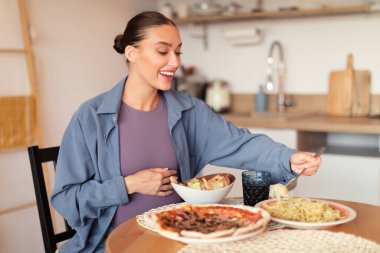 Pizza, salata ve makarna seçen, abur cubur arzulayan, mutfak masasında oturan beyaz bir kadın.