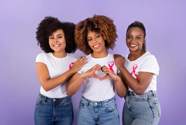 Oncology Awareness. Trio Of Positive African American Ladies With Pink Breast Cancer Ribbons Hugging, Woman Shaping Fingers Like Heart Over Purple Studio Background. Female Support Group