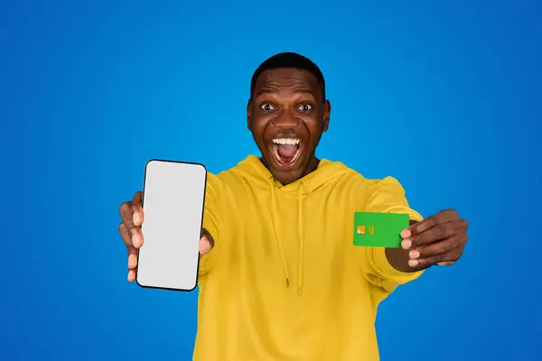 高兴的黑人年轻人展示了信用卡 带着空屏幕的电话 喜庆胜利 庆祝成功 在蓝色背景下被隔离 工作室 广告和提供 盈利和购物 — 图库照片