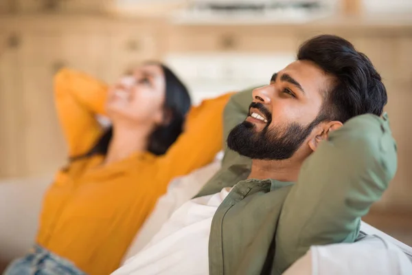 Time to relax. Calm indian couple resting together on comfortable couch at home, leaning back on sofa with hands behind head, selective focus on smiling man