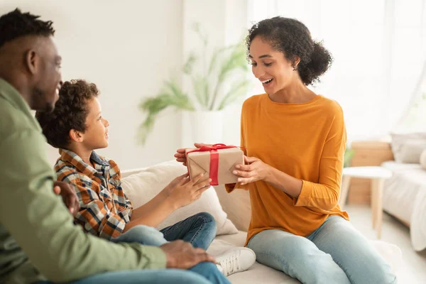 Family birthday celebration and presents. Young black father and son greeting surprised mother on holiday, boy giving wrapped festive gift box sitting together on couch at home