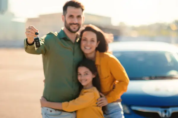 Excited family of three showing new car key, standing near luxury auto outdoors and smiling at camera, selective focus. Parents and daughter celebrating buying vehicle