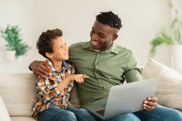 快乐的年轻非洲裔美国人的父亲和儿子坐在沙发上的笔记本电脑 浏览互联网 在家里看有趣的视频 男孩和爸爸一起购物时指指点点小玩意 — 图库照片