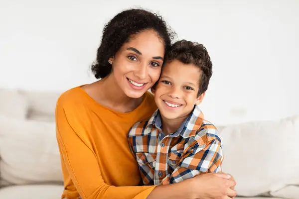 Portrait Of Happy Young Mom Embracing Her Kid Son Smiling To Camera At Home Interior. Boy Enjoying Mothers Hug Sitting On Couch Indoors. Family Bonding And Motherhood Concept