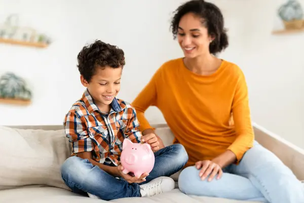Family Savings. Happy Little Boy And His Mom Holding Piggybank, Putting Coin At Home. Young Middle Eastern Mother Teaching Son Economy. Kid Personal Savings Concept. Selective Focus