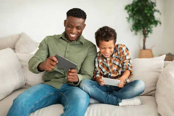 快乐的黑人爸爸和他的儿子在他们的智能手机上玩网络游戏 周末玩得开心 坐在现代家居的沙发上 家庭数码娱乐及小工具 — 图库照片