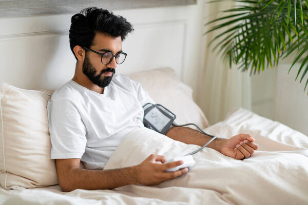 Young Indian Man Using Upper Arm Blood Pressure Monitor While Sitting In Bed, Millennial Eastern Guy Feeling Unwell, Measuring Arterial Tension At Home, Suffering Health Problems, Free Space