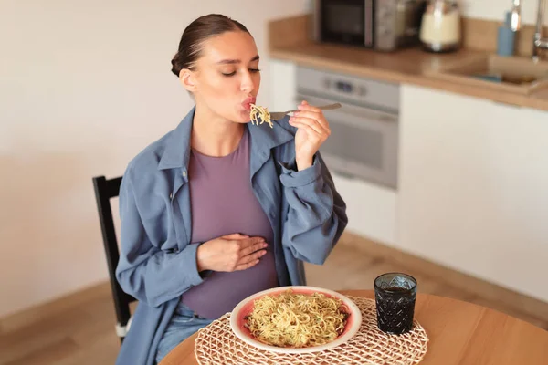 Hungry pregnant woman enjoy eating spaghetti, having desire for junk italian food, sitting at table in kitchen interior. Cheat meal, gluttony and fastfood concept