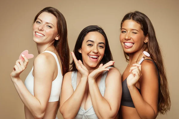 Glad millennial diverse women in underwear enjoy beauty care, use stone, roller from quartz, isolated on beige background, studio. Anti age, lifting treatment, dermatology and spa massage, wellness
