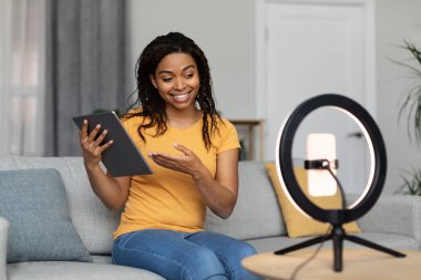 Modern iş ve hobi. Siyahi kadın blogcu video blogu çekiyor, tableti gösteriyor, oturma odasının içinde yayın yapıyor. Modern teknolojiyi inceleyin, reklam ve sunum