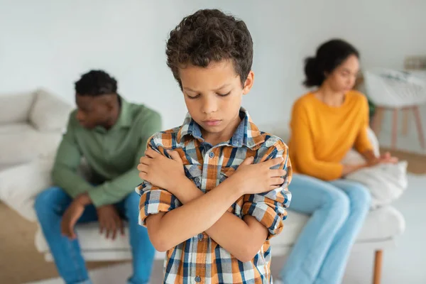 Divorce And Family Conflicts. Upset Son Crossing Hands While His Unhappy Multiracial Parents Sulking After Quarrel In The Background At Home, Portrait Of Depressed Kid Feeling Lonely