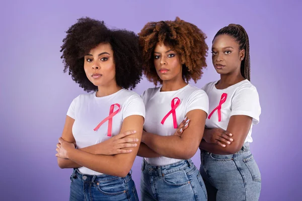 Breast Cancer Awareness Month. Three Serious African American Women In T-Shirts With Pink Ribbons Posing Together, Standing And Crossing Hands Over Purple Studio Background, Looking At Camera