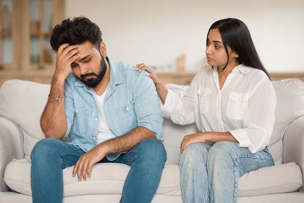 Crisis in marriage, divorce, breakup. Loving indian woman talking and touching her indifferent husband, couple sitting on couch at home, having quarrel