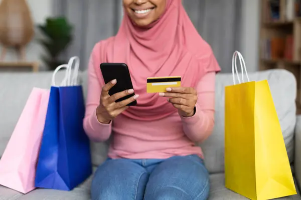 若いイスラム教徒の女性ショップアホリックは スマートフォンや銀行カードを使用して カラフルなショッピングバッグの間にソファーに座っています Eコマース 小売コンセプト — ストック写真
