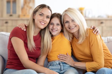 Mutlu sarışın güzel anne, büyükanne ve çocuk reşit olmayan kızın aile portresi. Sevimli bir kız çocuğu. Annesiyle büyükannesine sarılıyor. Aile fotoğrafı için kanepede poz veriyor.