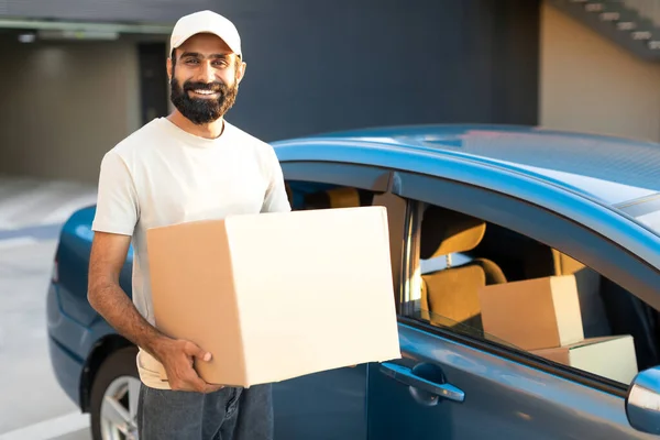 Parcels Shipping By Car. Middle Eastern Deliveryman Holds Cardboard Box Near His Modern Automobile, Standing Outside And Smiling At Camera, Advertising Great Delivery Offer
