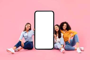 Mutlu üç çok ırklı bayan arkadaş beyaz ekranlı dev bir cep telefonunun yanında oturmuş tasarım, kolaj, taklit, ücretsiz alan sunuyorlar.