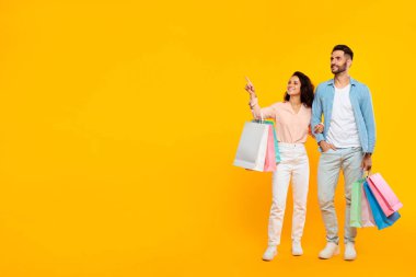 Mevsimlik indirimler. Alışveriş torbalarıyla ayakta duran mutlu eşler ve boş alana işaret eden, sarı arka planda poz veren Avrupalı çift, alışveriş anlaşmalarından hoşlanıyor, kopyalama alanı