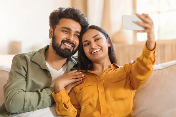 Loving indian couple sitting on sofa, embracing and taking selfie on cellphone, looking at phone screen and smiling, free space, home interior. Relationships concept