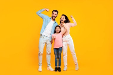 Aile gücü. Mutlu Avrupalı anne ve baba ve kız pazı kaslarını gösteriyor ve kameraya gülümsüyor, sarı stüdyo arka planında birlikte poz veriyorlar, tam boy