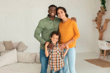 Mutlu anne babalar, oğullarını kucaklıyor ve kamera karşısında gülümsüyor. Modern oturma odasında bir arada duran ve sıcak bir kucaklaşmayı paylaşan üç kişilik aile.