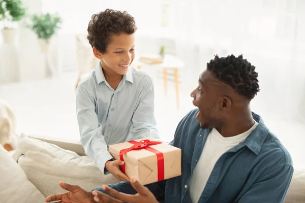 Fathers Day Surprise. Cute Little Boy Presenting Gift To His Black Dad, Surprising Joyful Daddy With Present In Wrapped Box, Celebrating Birthday Or Family Holiday And Bonding Together At Home