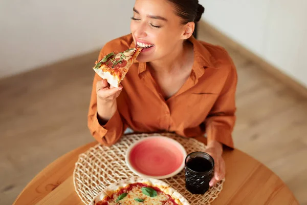 Excited woman enjoying eating tasty pizza, holding and biting slice, sitting at table in kitchen interior, free space, above view. Cheat meal, nutrition and fast food