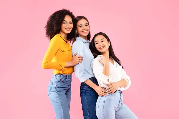 Diverse female friendship. Happy young multiracial ladies embracing and posing, smiling at camera, friends standing over pink studio background, free space
