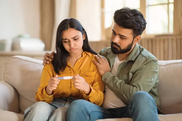 Infertility. Unhappy young indian couple holding negative pregnancy test result, sitting on sofa, man supporting wife in problem. Reproduction, childbirth healthcare struggle
