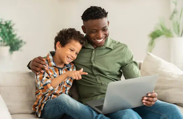 Happy Black Man With His Little Son Using Laptop Together At Home, Kid Boy Pointing At Computer Websurfing With Daddy Sitting On Sofa In Living Room. Gadgets And Family Online Fun