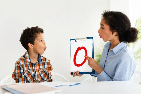 Speech Therapy, Speaking Lesson. School boy learning letter O with private tutor lady sitting at table indoor, teacher teaching kid pronunciation, curing childs problems and impediments