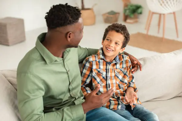 Cheerful African American Dad Talking To Little Son, Embracing And Sharing Funny Stories At Home. Kid Boy Enjoying Quality Time with Daddy On Weekend. Family Communication. Selective Focus