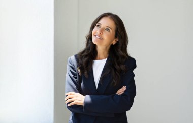 50 'li yaşlarda neşeli, çekici, olgun bir kadının portresi. Resmi takım elbiseli, kollarını göğsünde kavuşturarak ofiste poz veren ve fotokopi odasına bakan bir kadın. İş yerindeki mutlu iş kadınının fotoğrafı.