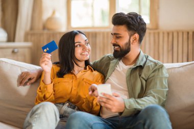 İnternetten aile alışverişi. Cep telefonu ve kredi kartı tutan neşeli Hintli eşler, mobil bankacılık uygulaması kullanarak, evdeki oturma odasında kanepede oturuyor.