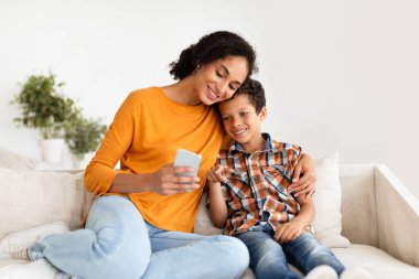 Mutlu anne ve küçük oğlu akıllı telefonları koltukta kucaklaşırken, uygulamayı kullanırken ve sosyal medyayı iç mekanda birlikte kaydırırken. Neşeli İki Kişilik İnternet Sörfçüsü Telefon Gadget ile