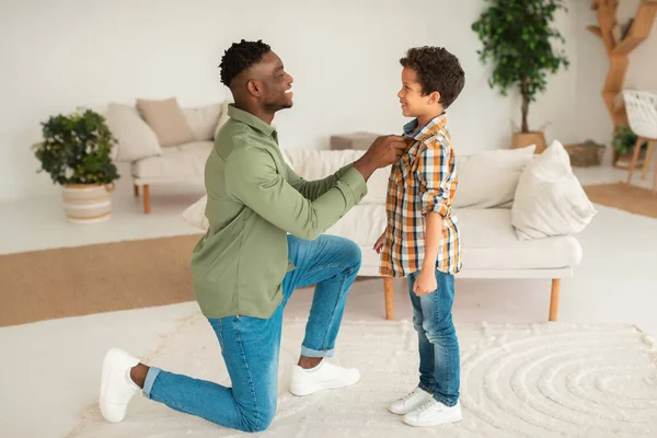 Black Young Father Getting His Kid Son Ready For School Day, Buttoning Shirt In Modern Living Room, Preparing Little Boy And Caring For His Neat Appearance At Home. Children Style And Fashion