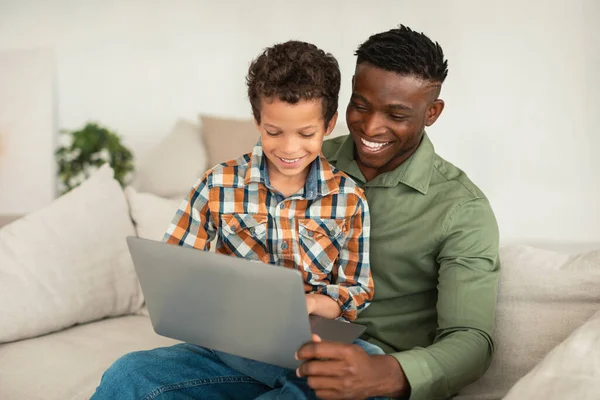 快乐的黑人爸爸坐在家里的沙发上 帮小儿子在笔记本电脑上上网 非裔美国人的父亲和孩子一起玩网络游戏或享受电子学习 — 图库照片