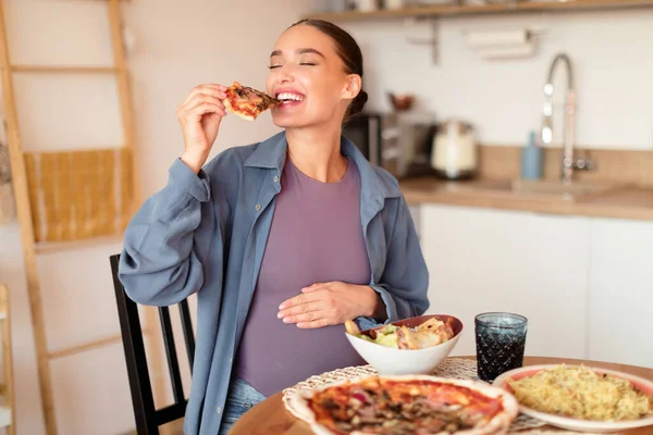 Young hungry pregnant woman biting slice of pizza, having desire for junk food, sitting at table full of dishes in kitchen interior. Cheat meal and fastfood concept