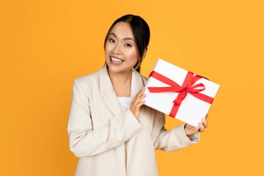 Takım elbiseli mutlu genç Asyalı kadın, sarı stüdyo arka planında izole edilmiş hediye kutusu gösteriyor. Halk duyguları, mevcut fikir, bayram kutlaması, doğum günü kutlaması, reklam ve teklif